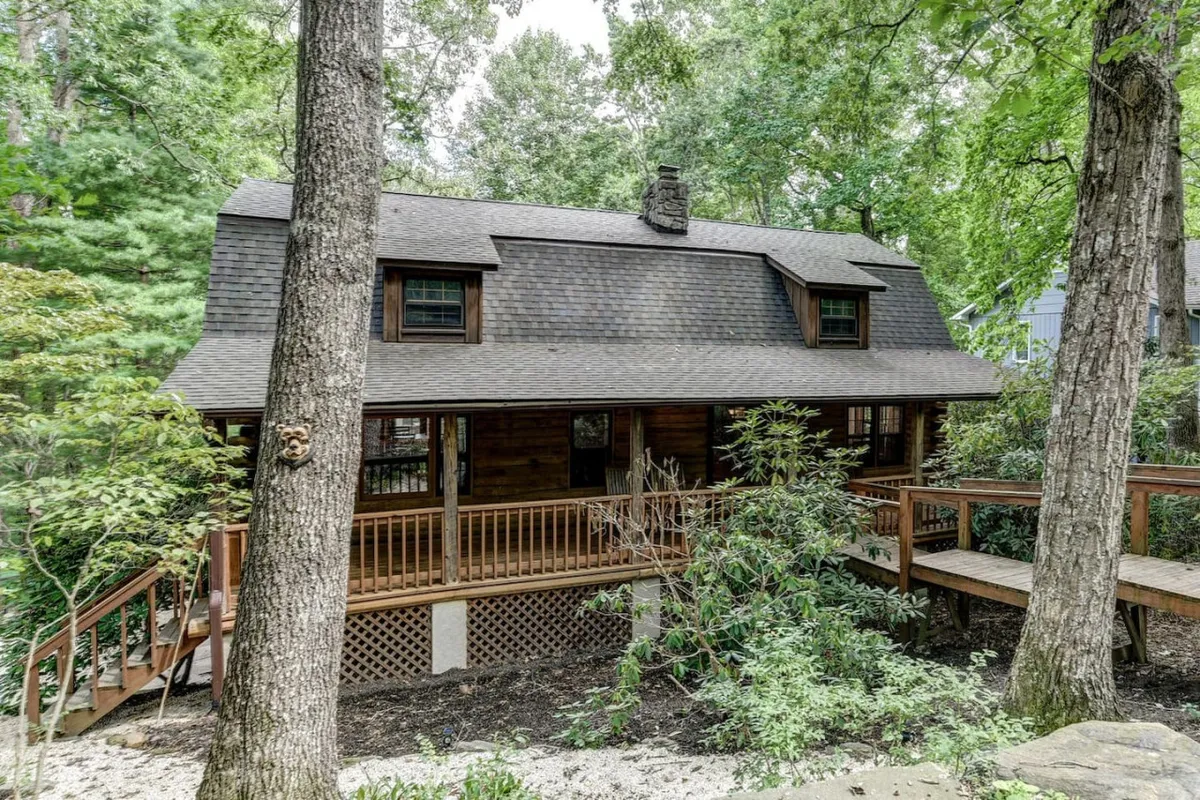 A tranquil scene featuring a spacious wooden deck surrounded by mature trees, with the cabin's shingled roof visible. The setting evokes opportunities for enjoying the outdoors, from sipping coffee in the morning to hosting relaxed gatherings with friends or watching the sunset.