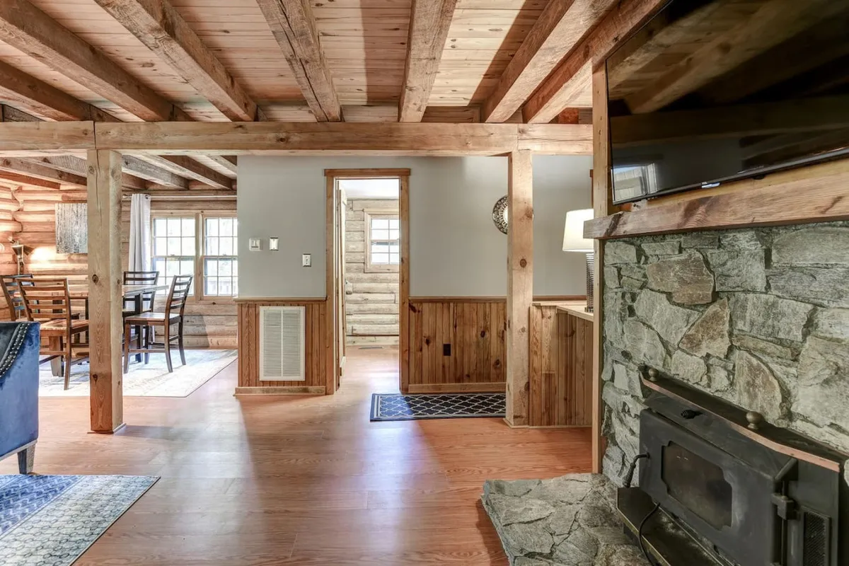 A welcoming interior scene centered on a stone fireplace flanked by wooden beams, with a dining table and a blue accent chair nearby. The space exudes warmth and hospitality, suggesting a place for family meals, board games, or storytelling by the fire. The combination of natural textures and soft furnishings draws people in for communal experiences.