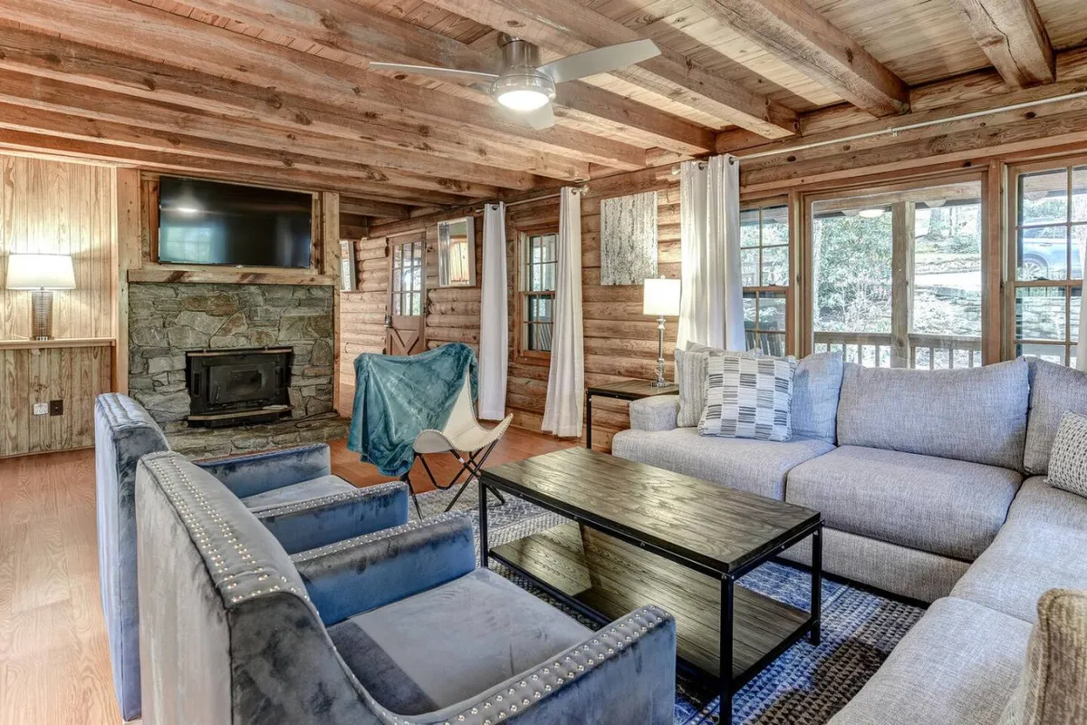 The image showcases a warm, inviting living area with a rustic stone fireplace as the centerpiece, surrounded by plush, comfortable seating. Large windows flood the space with natural light, highlighting the rich wooden beams and cozy cabin ambiance—perfect for relaxing, socializing, or enjoying quality family time.