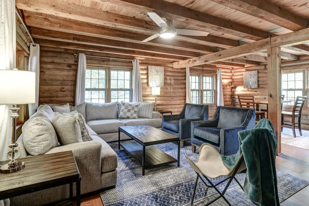 A spacious and inviting living area featuring a plush sofa, two armchairs, a sturdy coffee table, and rustic wooden beams overhead. Natural light pours in from the windows, illuminating the warm textures and cozy furnishings that define the space. The layout encourages conversation and shared activities, with ample room for board games or quiet relaxation.