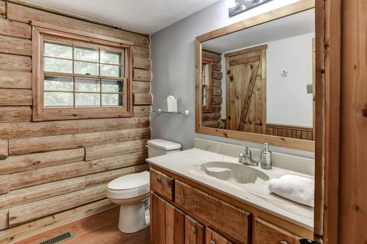 A warmly inviting shared bathroom featuring wooden log walls, a vanity with a sink and large mirror, a window bringing in natural light, and a clean, uncluttered layout. The space is practical yet aesthetically consistent with the upstairs retreat vibe.