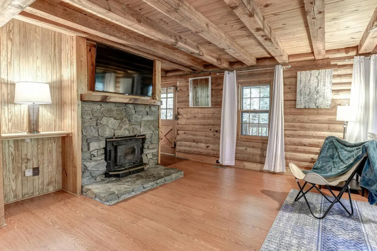 A welcoming living space featuring a stone fireplace, exposed wooden beams, and large windows that offer glimpses of the tranquil outdoors. Comfortable soft furnishings invite relaxation, and the room is bathed in natural light, further blurring the line between interior comfort and the surrounding natural beauty.