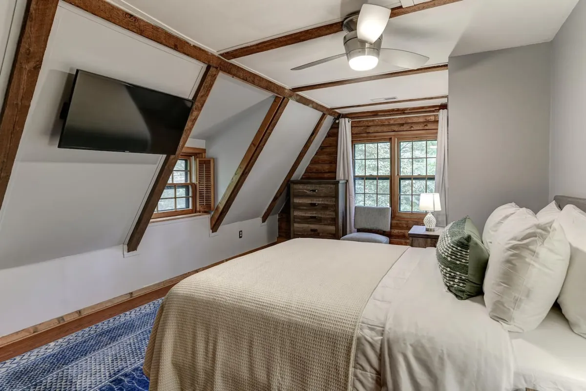 A serene king bedroom featuring textured bedding, exposed wooden ceiling beams, a large window filling the space with natural light, a dresser, and an inviting armchair. The tranquil setting is ideal for relaxation and privacy, echoing the restful atmosphere described in the section.