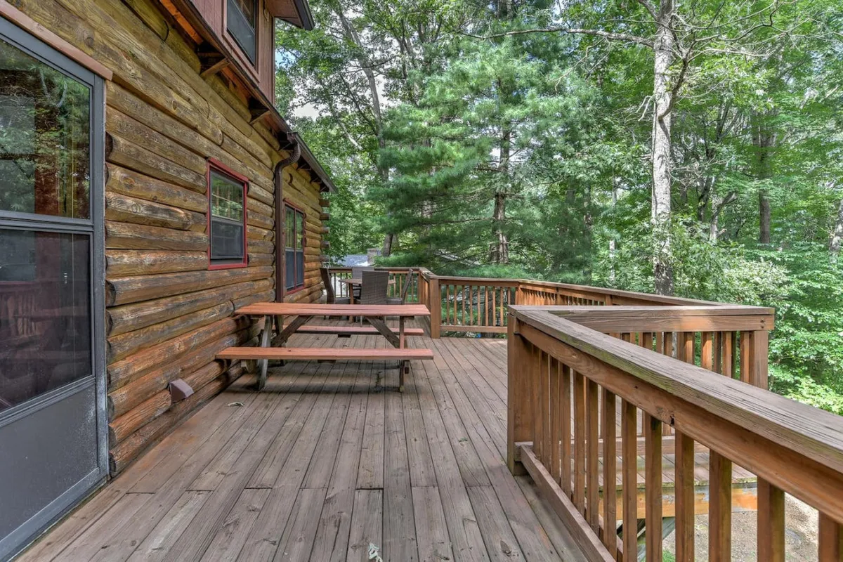 A spacious wooden deck nestled among tall trees, with sturdy outdoor seating arranged for gatherings. The log cabin structure provides a rustic backdrop, while the view hints at the tranquility of the surrounding nature, perfect for evening conversations or stargazing around a fire pit.