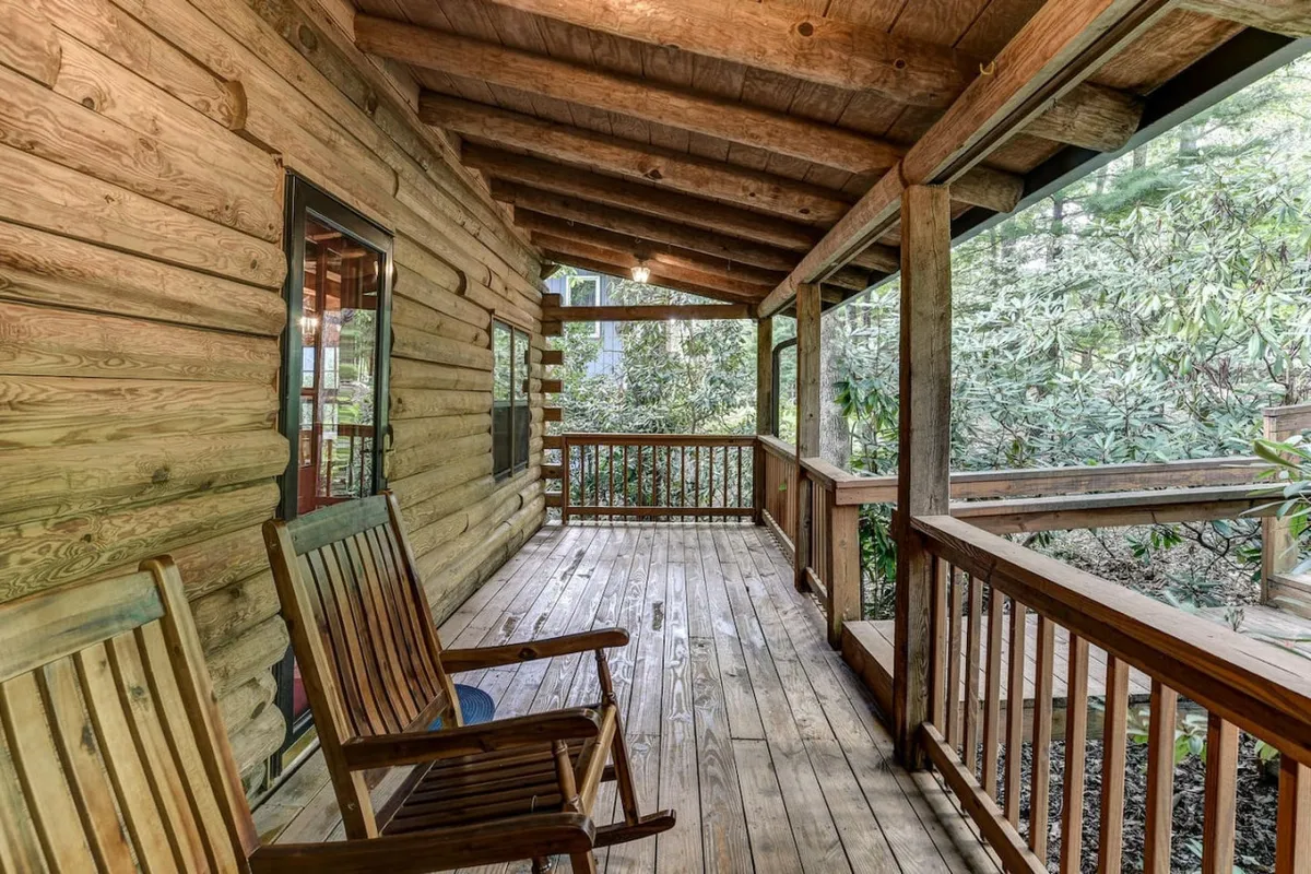 A close-up view of the log cabin’s porch with rocking chairs, set against a vibrant green backdrop. The scene suggests quiet contemplation, watching sunrises or sunsets, and gatherings in a natural setting.