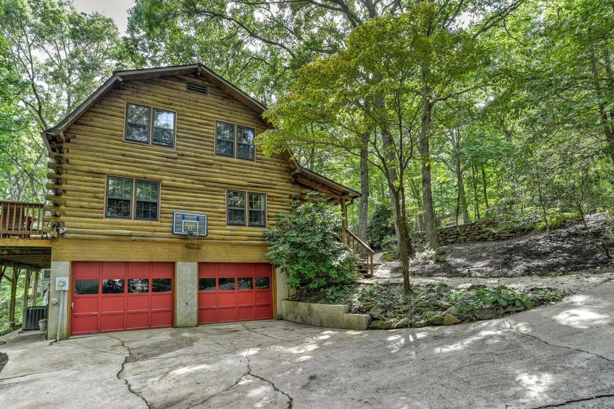 The image showcases the log cabin structure with red garage doors, framed by lush greenery. The open outdoor space suggests areas for kids to play, family gatherings, and stargazing, making it visually clear that the property is well-suited for family activities.