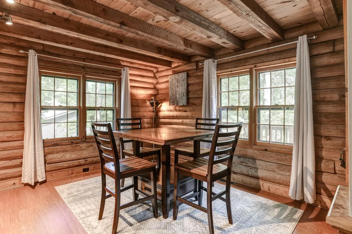 A rustic dining area with a solid wooden table and four black chairs, set against wooden walls adorned with decorative art. The large windows let in streams of daylight and offer verdant views, creating a seamless transition between indoor dining and the natural world outside.