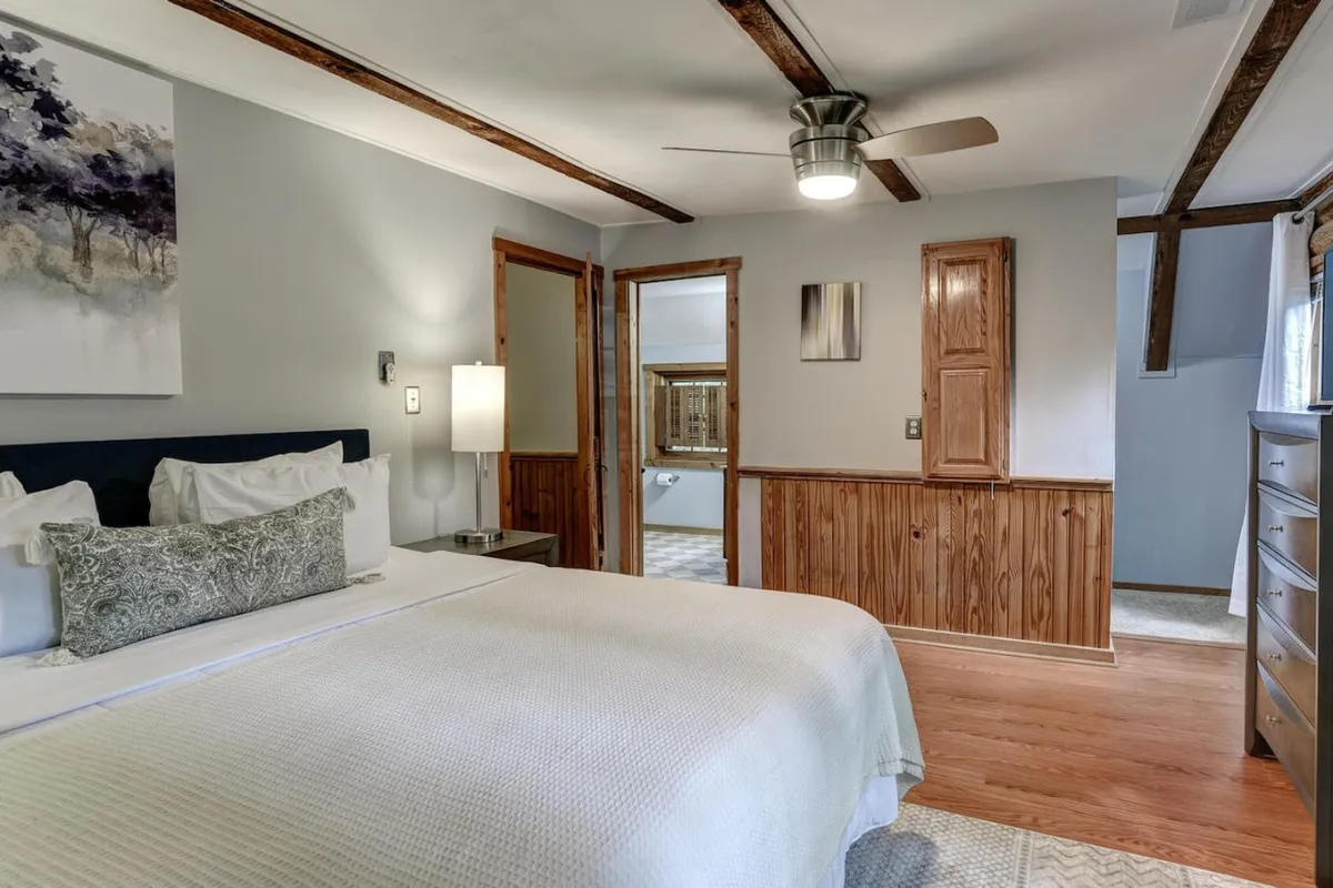 A calm and stylish queen bedroom with a bed adorned with decorative pillows, warm wood accents, an art piece above the bed, and a dedicated dressing area with mirrors. The room is airy and bright, evoking a sense of peaceful retreat.