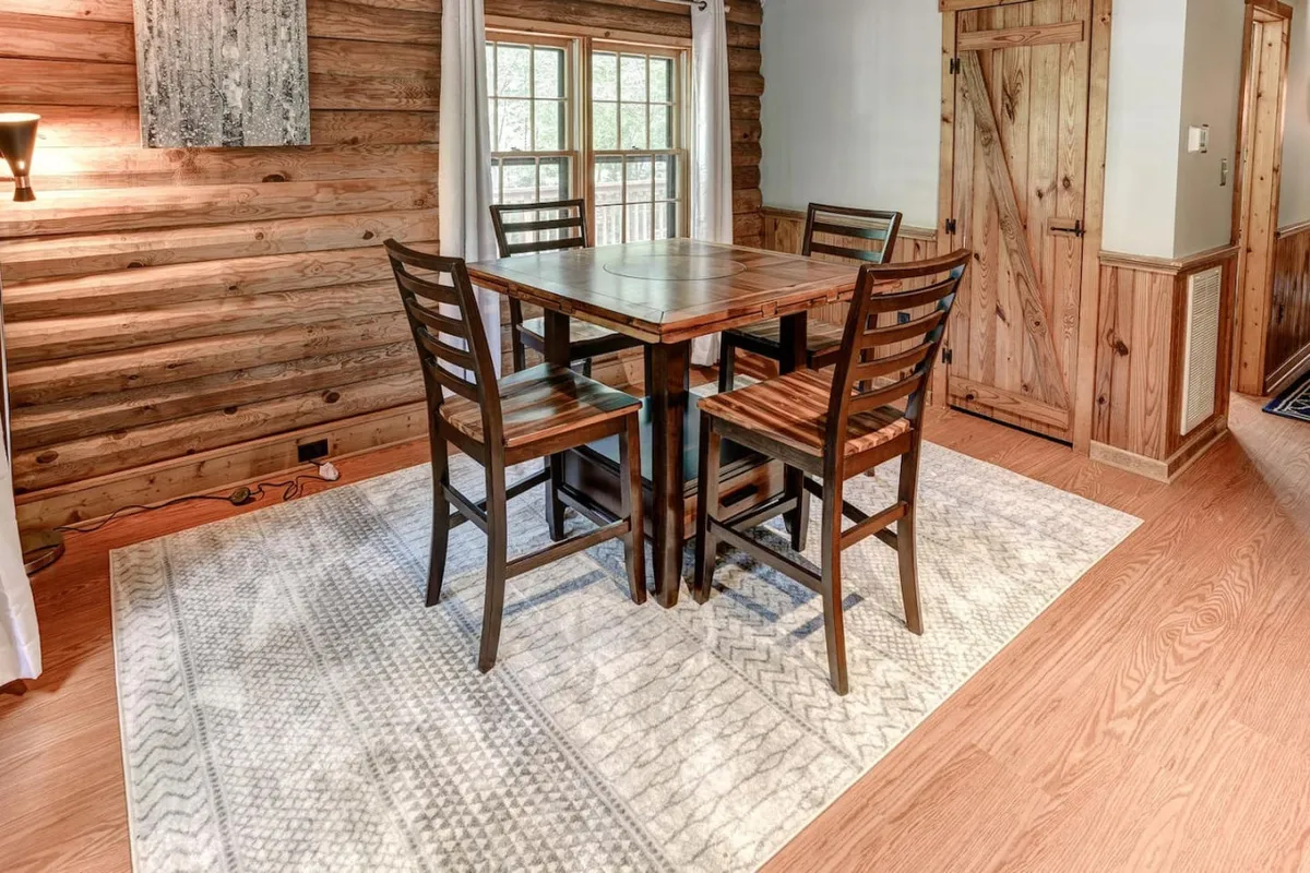 An intimate view of a wooden dining table surrounded by high-backed chairs, atop a textured area rug, with sunlight filtering through a nearby window. The setup radiates warmth and invites family and friends to sit down for meals or engage in casual game nights, underscoring the cabin's focus on togetherness.